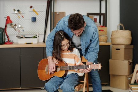 Photo for Young man in denim shirt and blue jeans bending over his daughter with guitar sitting on chair in garage during lesson of music at leisure - Royalty Free Image