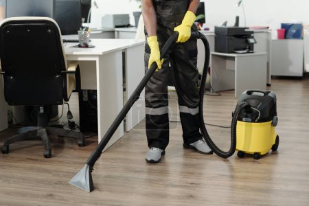 Téléchargez les photos : Jeune homme noir en uniforme et gants jaunes utilisant un aspirateur pour nettoyer le sol d'un grand bureau contemporain à aire ouverte - en image libre de droit