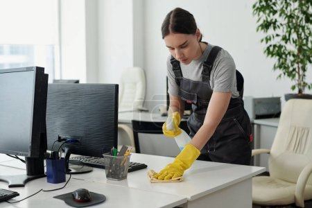 Foto de Young female staff of cleaning service company with plastic bottle of detergent wiping desk with duster in openspace office - Imagen libre de derechos