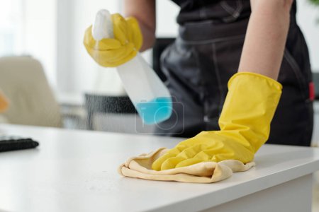 Photo for Young woman in protective yellow rubber gloves wiping dust on desk while spraying detergent and cleaning furniture in office - Royalty Free Image