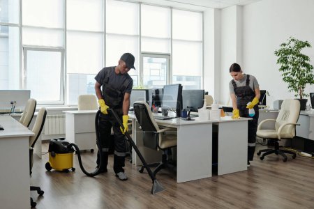 Photo for Young black man in workwear cleaning floor while girl wiping desks with computer monitors in large contemporary openspace office - Royalty Free Image