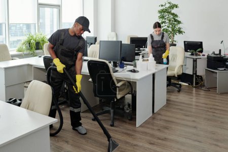 Téléchargez les photos : Two young workers of contemporary cleaning service company in coveralls and gloves carrying out their work in openspace office - en image libre de droit