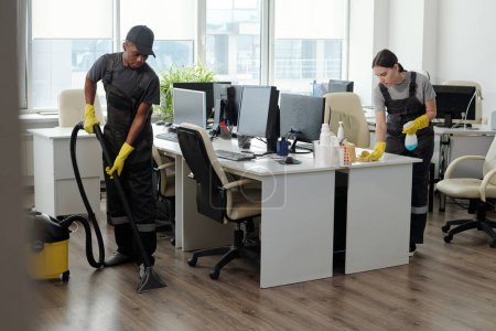 Foto de Young male and female workers of service company using detergent and vacuum cleaner for cleaning contemporary openspace office - Imagen libre de derechos