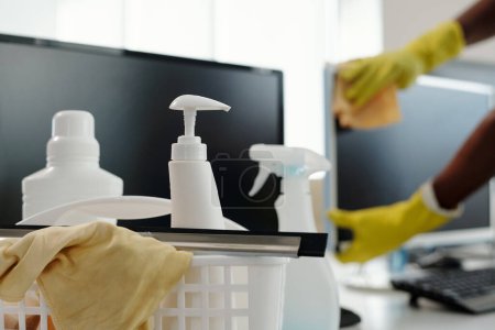 Photo for Group of detergents in white plastic bottles with dispensers and duster in basket against computer screen and gloved hands of man - Royalty Free Image