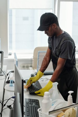 Photo for African American male staff of cleaning company in black t-shirt, cap and coveralls and yellow gloves wiping computer keyboard in office - Royalty Free Image