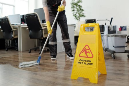Photo for Yellow plastic signboard with caution warning about wet floor and female cleaner in black uniform working with mop in openspace office - Royalty Free Image