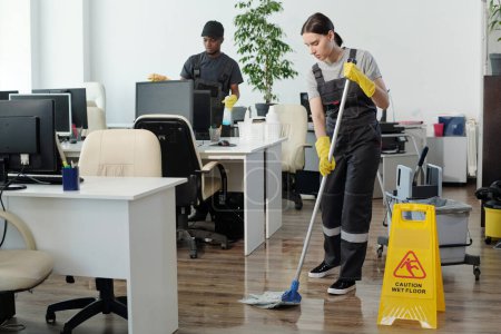 Foto de Young black man wiping computer monitors while Caucasian woman in coveralls and yellow gloves with mop cleaning floor in office - Imagen libre de derechos