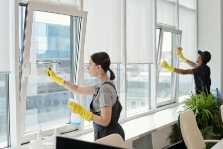 Photo for Young female cleaner in workwear washing large windows of openspace office against her African American male colleague - Royalty Free Image