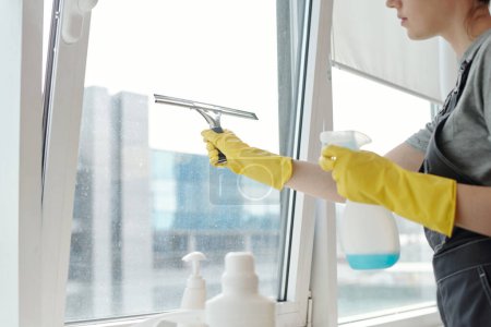Photo for Hands of young female cleaner in yellow gloves and workwear washing large office windows with detergent and silicone spatula - Royalty Free Image