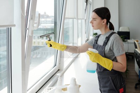 Photo for Young female staff of cleaning company washing large office window with special silicone spatula and detergent in plastic bottle - Royalty Free Image