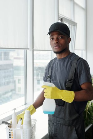 Foto de Contemporary young African American man with detergent in plastic bottle standing by sill with container while washing windows - Imagen libre de derechos