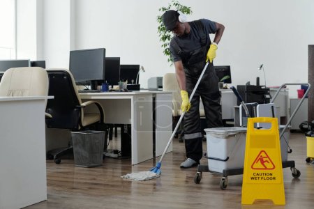 Joven hombre negro contemporáneo en el piso de limpieza de ropa de trabajo en la oficina de espacio abierto delante de letrero de plástico amarillo con precaución