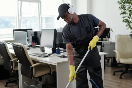 Photo for Young male staff of cleaning service company in headphones washing floor in large openspace office and listening to music - Royalty Free Image
