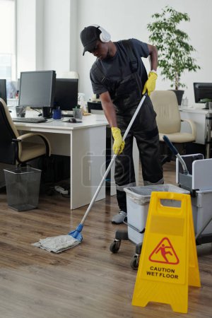 Foto de Young African American male janitor in headphones bending forwards while washing floor in large contemporary openspace office - Imagen libre de derechos