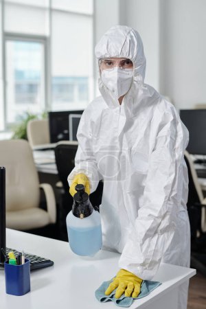 Photo for Female worker of cleaning service in protective workwear disinfecting desks and other surfaces in modern openspace office - Royalty Free Image