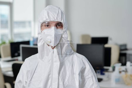 Photo for Young woman in white overalls, respirator and protective eyeglasses looking at camera while working in modern openspace office - Royalty Free Image