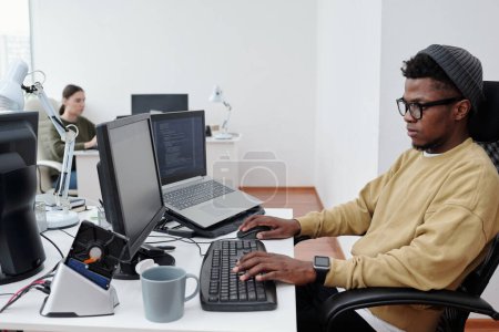 Téléchargez les photos : Jeune afro-américain il développeur en casual-wear décodage des données tout en étant assis devant l'écran d'ordinateur dans le bureau ouvert moderne - en image libre de droit