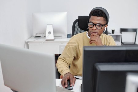 Téléchargez les photos : Jeune homme afro-américain sérieux en tenue décontractée et lunettes décodage des données devant les écrans d'ordinateur au bureau - en image libre de droit