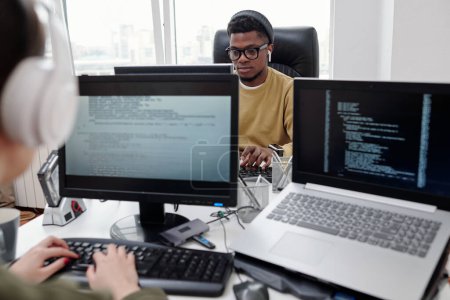 Téléchargez les photos : Jeune programmeur masculin sérieux regardant l'écran d'ordinateur pendant le réseau alors qu'il était assis devant sa collègue féminine au bureau - en image libre de droit