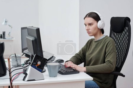 Téléchargez les photos : Jeune femme sérieuse en tenue décontractée écoutant de la musique dans des écouteurs assis sur le lieu de travail et décodant des données devant un ordinateur - en image libre de droit