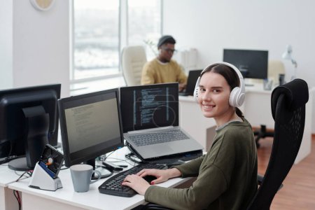 Photo for Happy young female it-engineer in headphones sitting by desk in front of computer monitor and laptop and looking at camera in office - Royalty Free Image