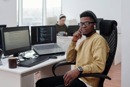 Téléchargez les photos : Jeune homme noir confiant en tenue décontractée avec écouteurs assis par le lieu de travail avec moniteur de calcul et ordinateur portable avec des données décodées - en image libre de droit