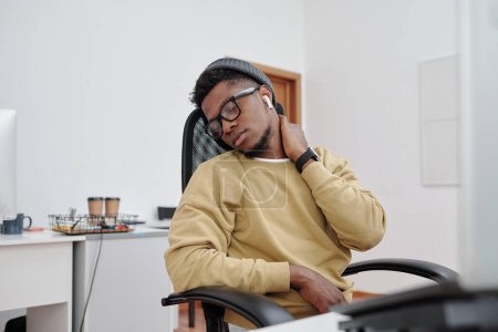 Téléchargez les photos : Young African American man in casualwear, eyeglasses and earphones doing relaxing exercise for neck by his workplace in office - en image libre de droit