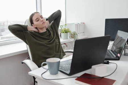 Foto de Young relaxed female programmer doing exercise for neck and arms while sitting by her workplace and looking at laptop screen - Imagen libre de derechos