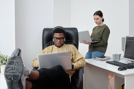 Téléchargez les photos : Jeunes développeurs interculturels masculins et féminins avec des ordinateurs portables travaillant individuellement dans le bureau tandis que l'un d'eux assis dans un fauteuil - en image libre de droit