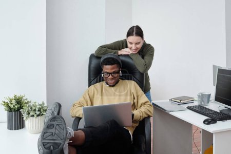 Photo for Two happy young multiracial software developers looking at screen of laptop on knees of African Americaan man sitting in armchair - Royalty Free Image