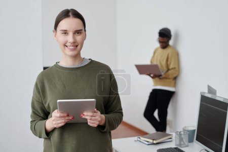 Téléchargez les photos : Young successful female programmer or manager with tablet looking at camera while standing against African American male colleague - en image libre de droit