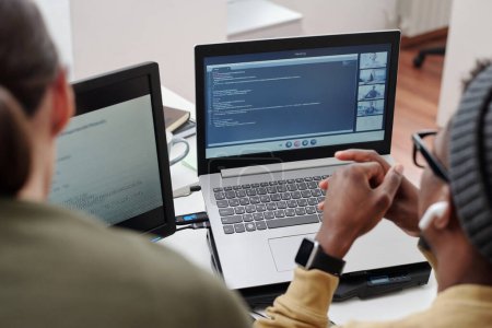 Téléchargez les photos : Mains de jeune homme noir sur le clavier de l'ordinateur portable avec chat vidéo à l'écran tout en étant assis par le lieu de travail à côté d'un collègue féminin - en image libre de droit