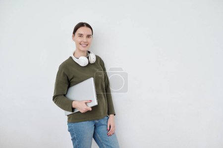 Photo for Young contemporary female student with folded laptop standing by white wall and looking at camera with copyspace on her left - Royalty Free Image