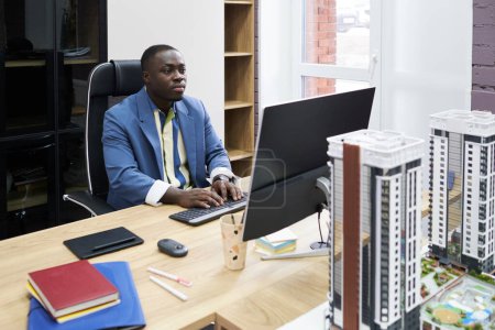 Téléchargez les photos : Architecte sérieux africain regardant moniteur tout en tapant sur l'ordinateur sur son lieu de travail au bureau avec des modèles de bâtiments sur la table - en image libre de droit