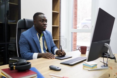 Téléchargez les photos : Sérieux concepteur africain regardant l'écran d'ordinateur tout en travaillant sur une tablette graphique à la table au bureau - en image libre de droit