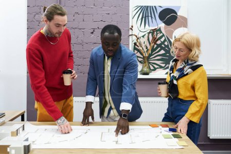 Photo for African man pointing at blueprint on table and discussing new project together with his colleagues - Royalty Free Image