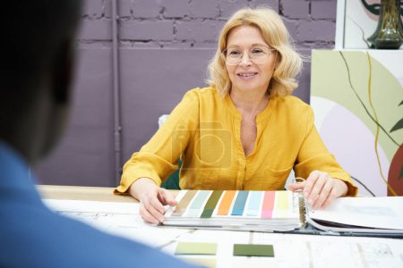 Photo for Mature interior designer sitting at table with palette and discussing colour with her client during meeting at office - Royalty Free Image