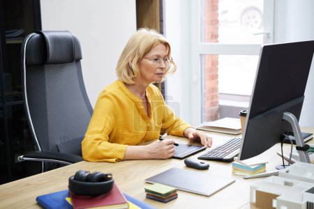 Téléchargez les photos : Femme sérieuse mature regardant l'écran d'ordinateur et travaillant sur une tablette graphique tout en étant assise à son lieu de travail au bureau - en image libre de droit
