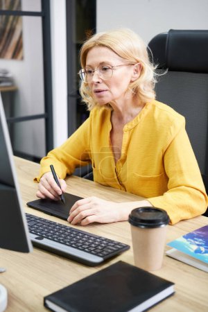 Foto de Serious mature woman in eyeglasses sitting at her workplace with computer using graphic tablet at her work - Imagen libre de derechos