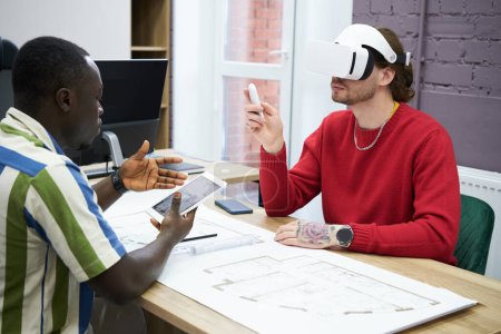 Foto de Business people sitting at table with blueprint using digital tablet to connect with VR goggles during teamwork at office - Imagen libre de derechos