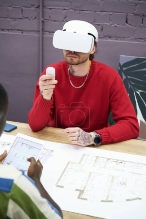 Photo for Young designer sitting at table with blueprint and using virtual reality headset to look at new project - Royalty Free Image
