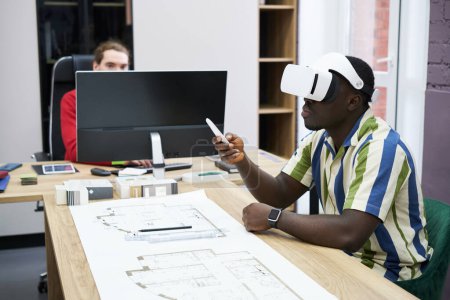 Photo for African young architect using virtual reality headset to look at construction project while sitting at table with blueprint - Royalty Free Image