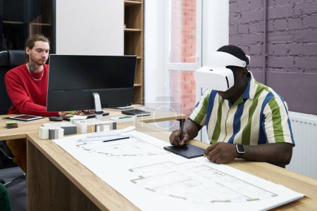 Photo for African designer in virtual reality goggles using graphic tablet at table with blueprint during work at office - Royalty Free Image