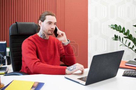 Foto de Young businessman sitting at office desk with laptop and having conversation on mobile phone - Imagen libre de derechos