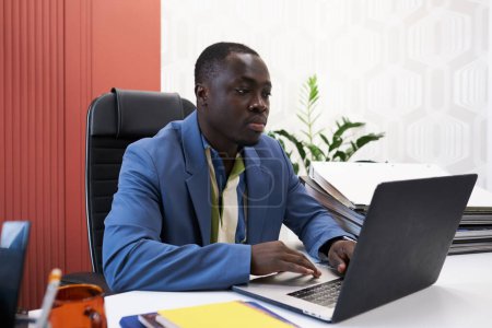 Téléchargez les photos : African male designer using his laptop to create new project while sitting at desk at office - en image libre de droit