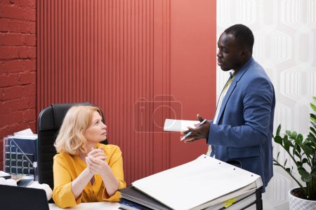 Foto de African businessman talking to businesswoman while she working at her workplace at office - Imagen libre de derechos