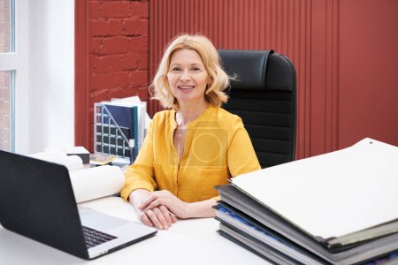 Foto de Retrato de mujer de negocios madura sonriendo a la cámara mientras está sentado en el escritorio con el ordenador portátil y pila de carpetas - Imagen libre de derechos