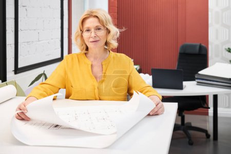 Foto de Portrait of mature female architect in eyeglasses looking at camera while working with blueprints of new buildings at table - Imagen libre de derechos