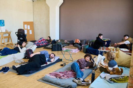 Foto de Varios refugiados descansando en sus lugares de dormir en el suelo de madera en una habitación espaciosa, mientras que la mujer joven habla con su hijo con libro - Imagen libre de derechos