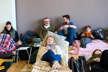 Foto de Mujer rubia joven usando tableta contra grupo de hombres hablando en camas mientras descansan en lugar de dormir preparado para migrantes - Imagen libre de derechos